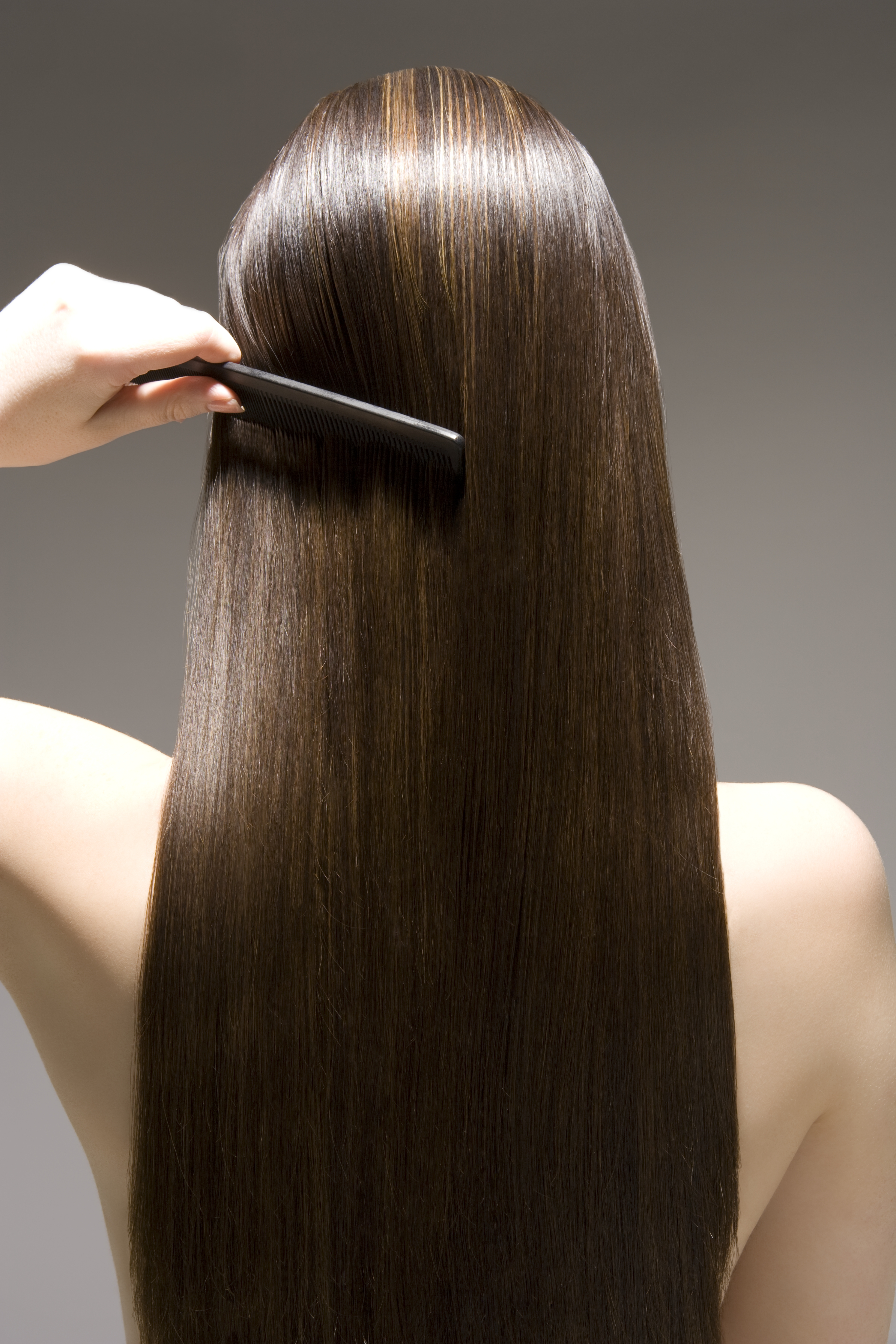 the back of a woman&#039;s head with long shiny brown hair. she&#039;s combing through it with a black comb