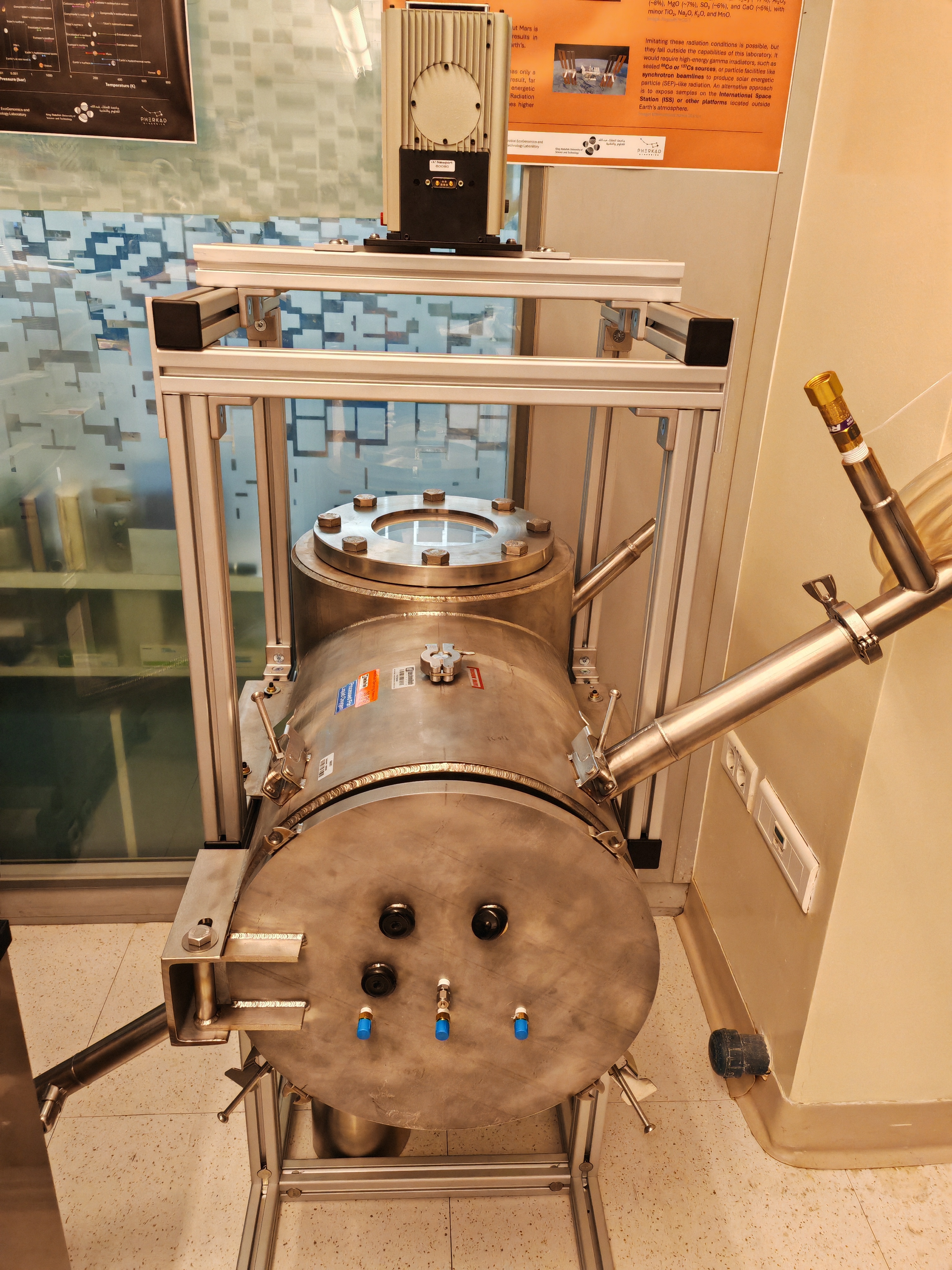 Photo of a large metal chamber about the size of a wine barrel, with several valves and gauges coming out of it, sitting on its side on a metal rack, inside a laboratory.