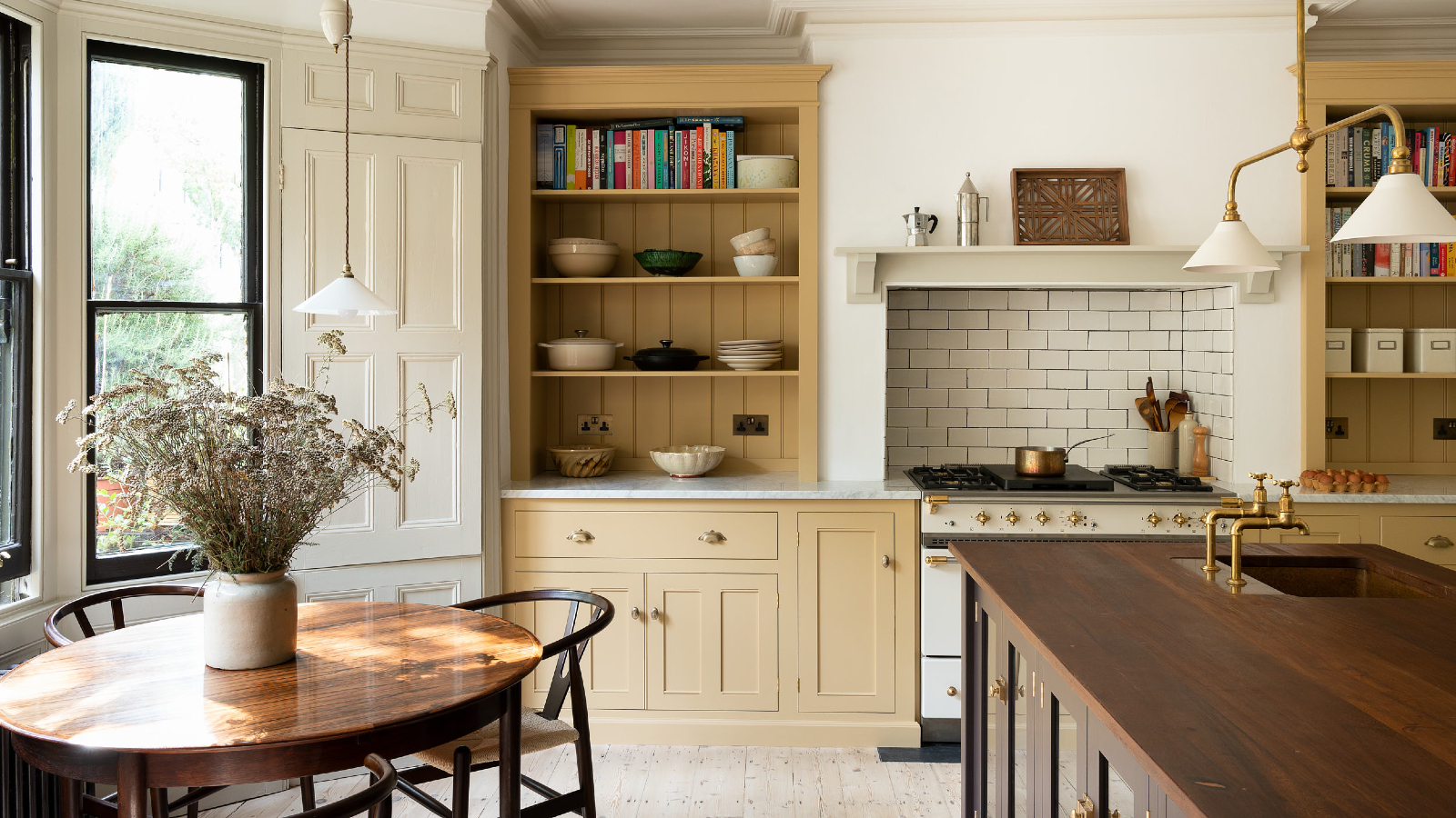 kitchen diner with yellow cabinetry and counter cabinets, metro tile backsplash behind stove, wooden kitchen island with sink, brass faucet, round mahogany table with black chairs, black painted window frames, enamel and brass pendant above island, wood floorboards
