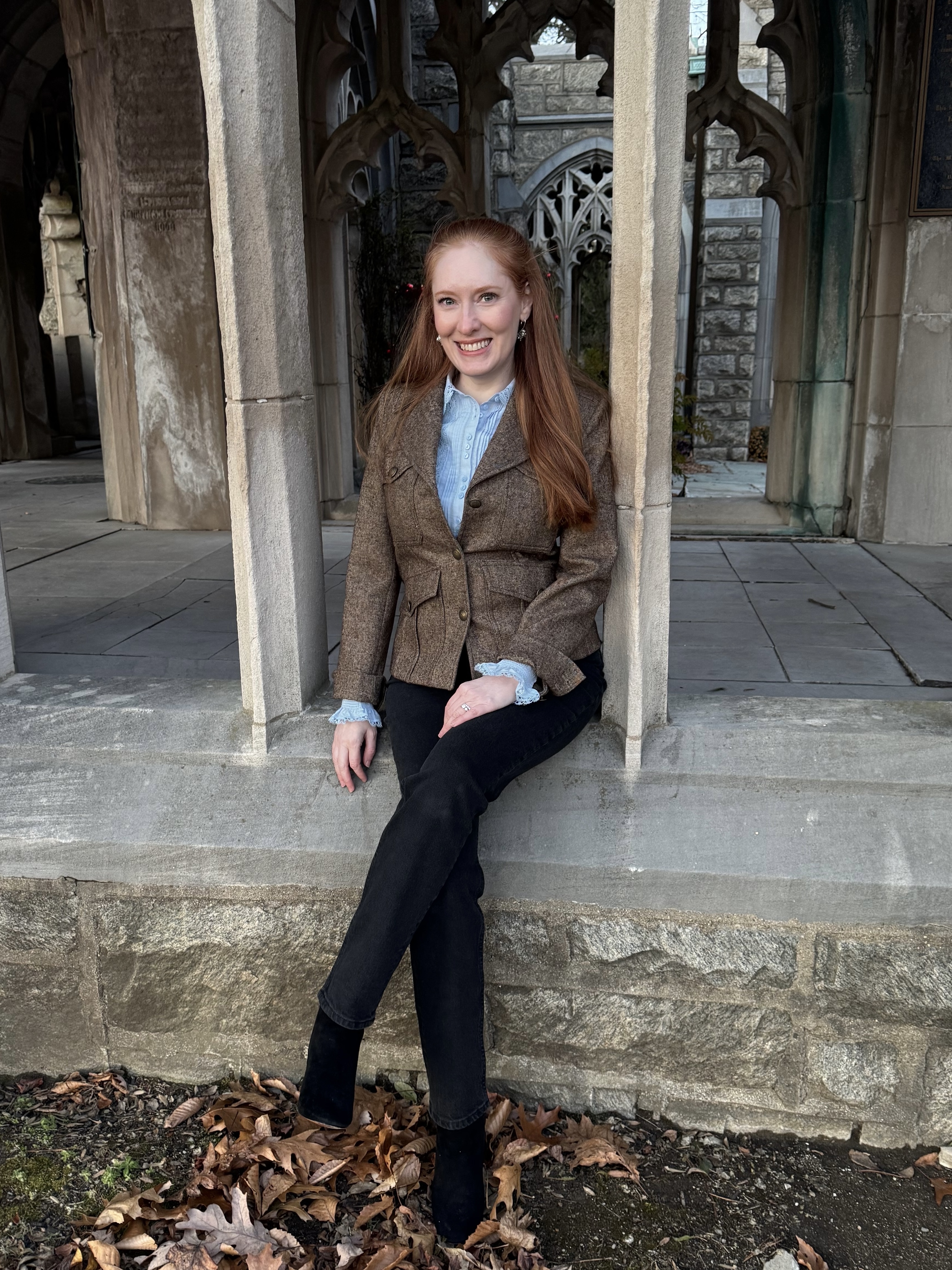 Kristin Contino sitting on a wall wearing a brown jacket, blue blouse and black jeans