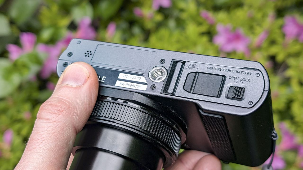 Close up of the Panasonic Lumix TZ300 compact camera's battery door with a colorful floral backdrop, view of the top and the lens is extended