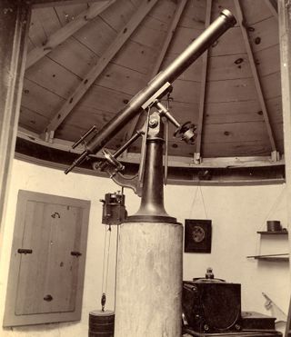 a black and white picture of a telescope mounted on a long pole. on the left is a wooden window with shutter closed. behind is a picture of the moon on the wall and various small shelves and objects