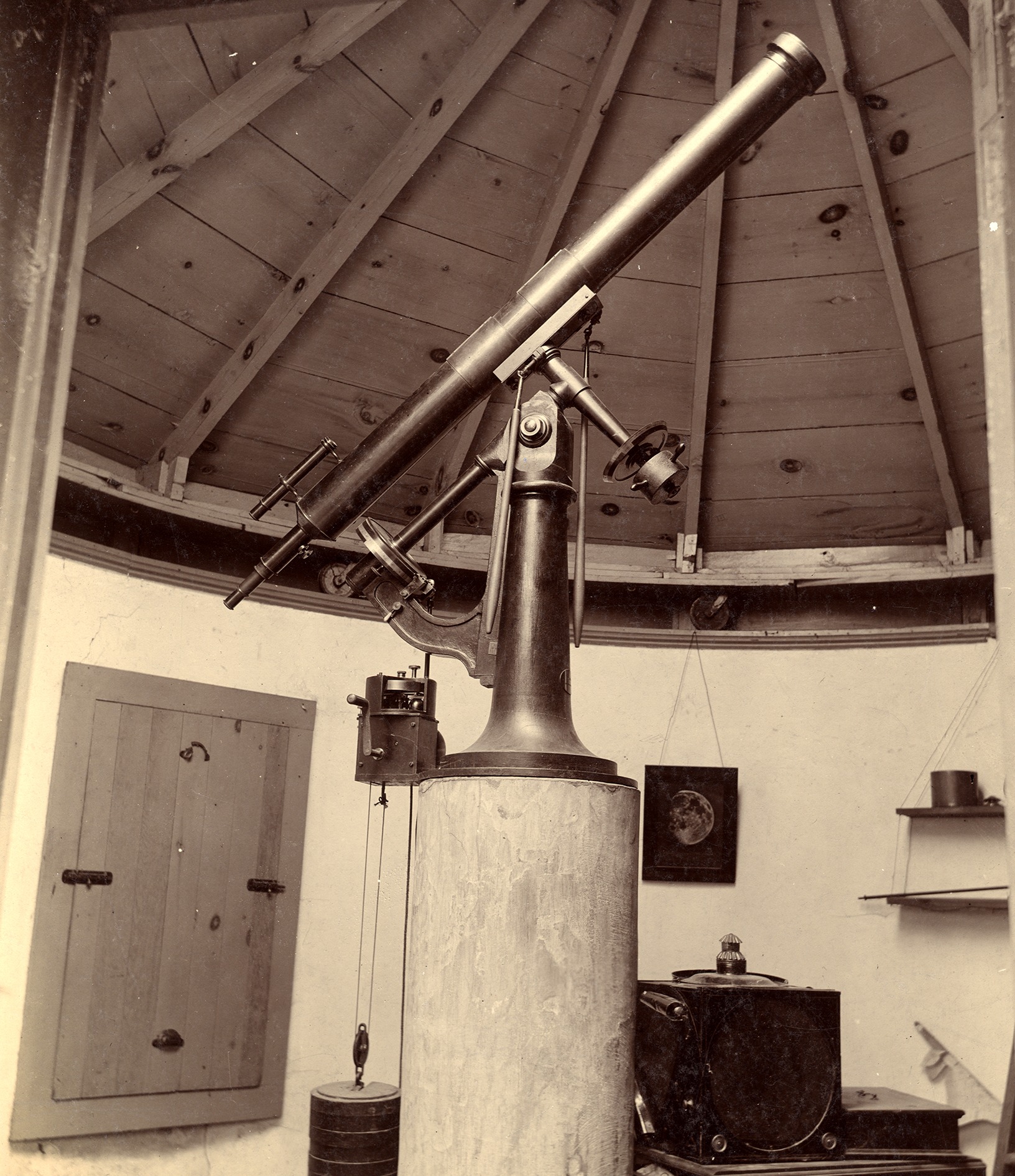 A black and white photograph of a telescope mounted on a long pole. On the left is a wooden window with shutters closed.The back wall has a moon painting and various small shelves and objects