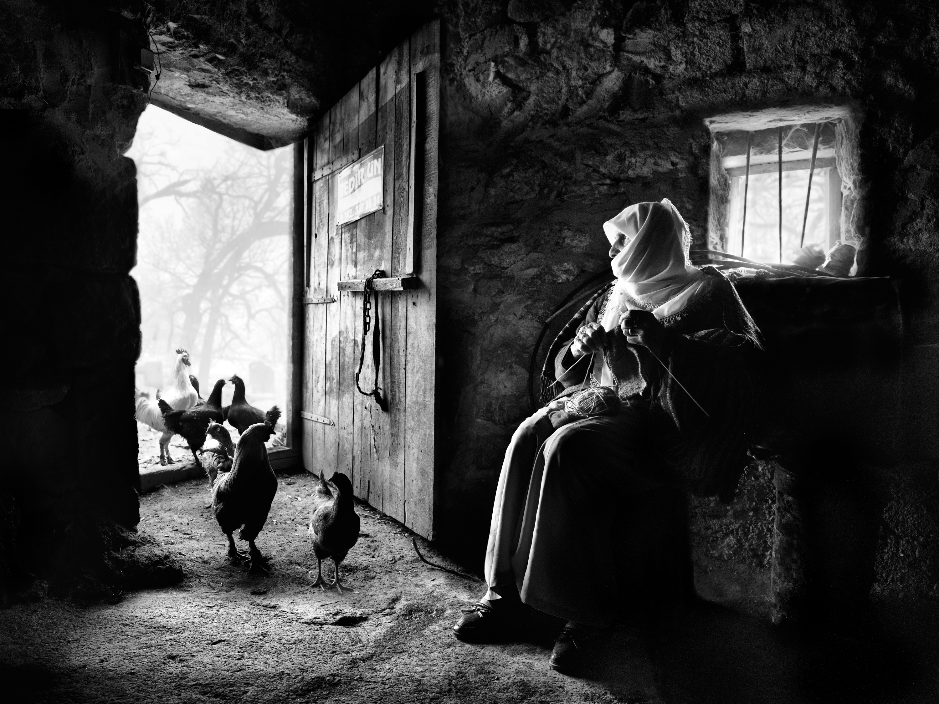 Black and white photograph of a woman in a headscarf sitting in a stone doorway knitting, with chickens gathered on the threshold and a padlocked wooden door beside her.