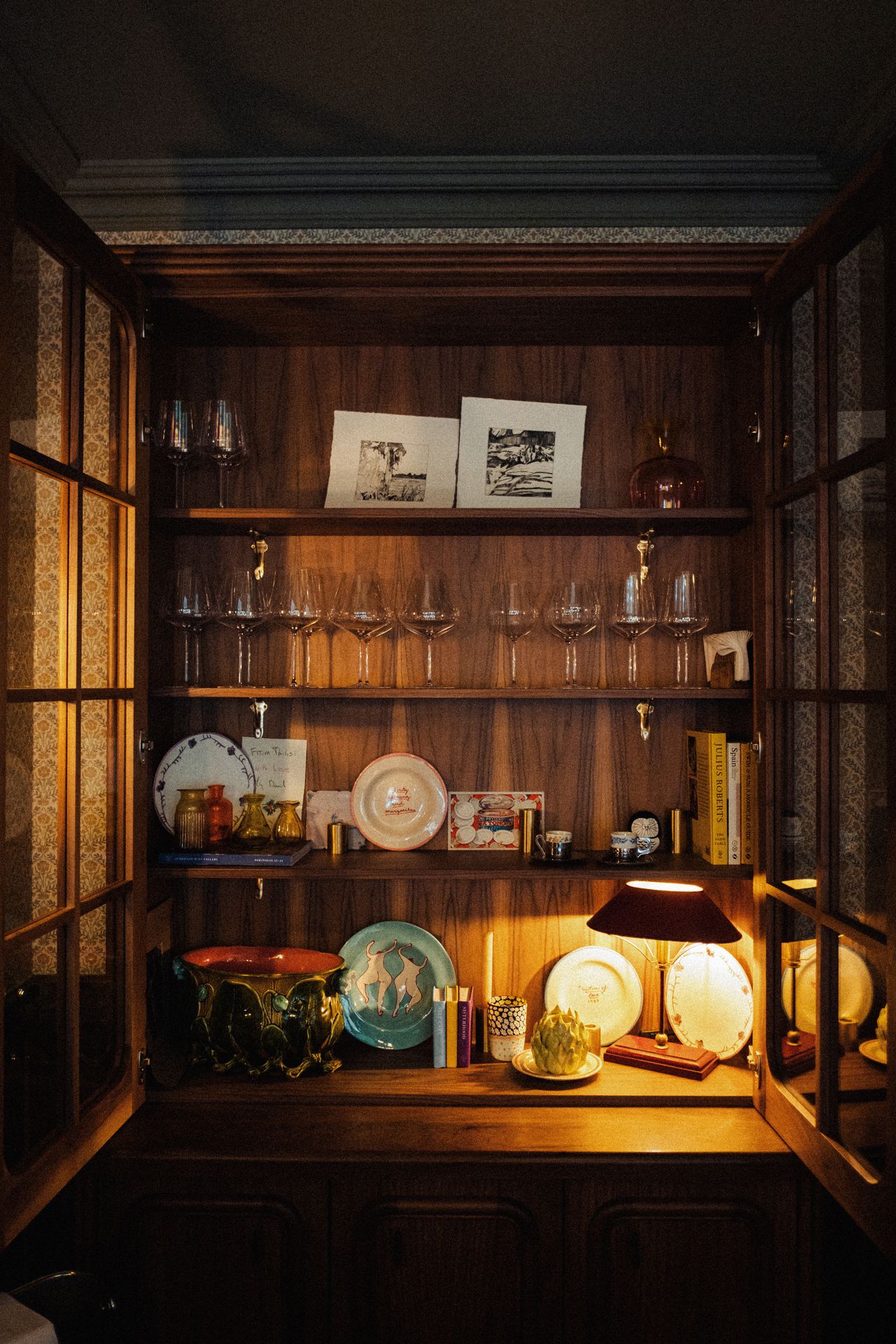 A wooden cabinet is filled with collectibles, including glasses, ceramics, and postcards, and brought to life by warm light.