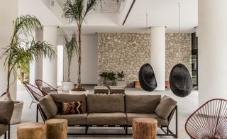 A sitting area with a brown sofa, round wooden coffee tables, a variety of different shaped chairs, stone walls, white pillars and potted plants.