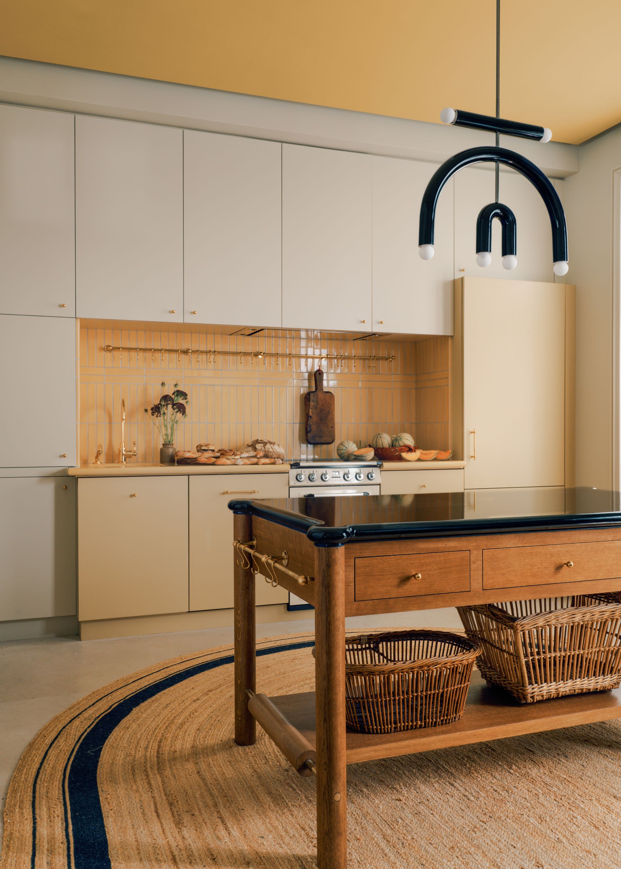 A modern kitchen with off-white cabinets and lower butter yellow cabinets. A dark yellow ceiling, a freestanding wood island unit, a jute rug, and a black modern sculptural pendant light.