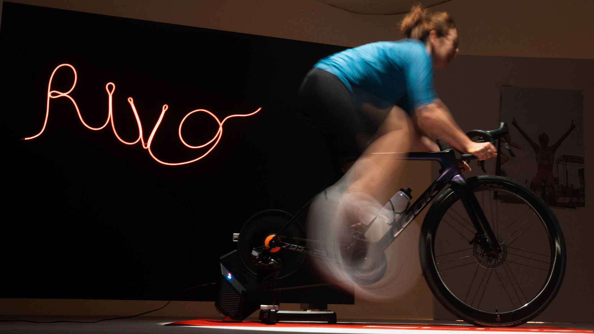 Lady riding an Elite Rivo with Zwift Cog and Click smart trainer with Rivo neon sign in the background