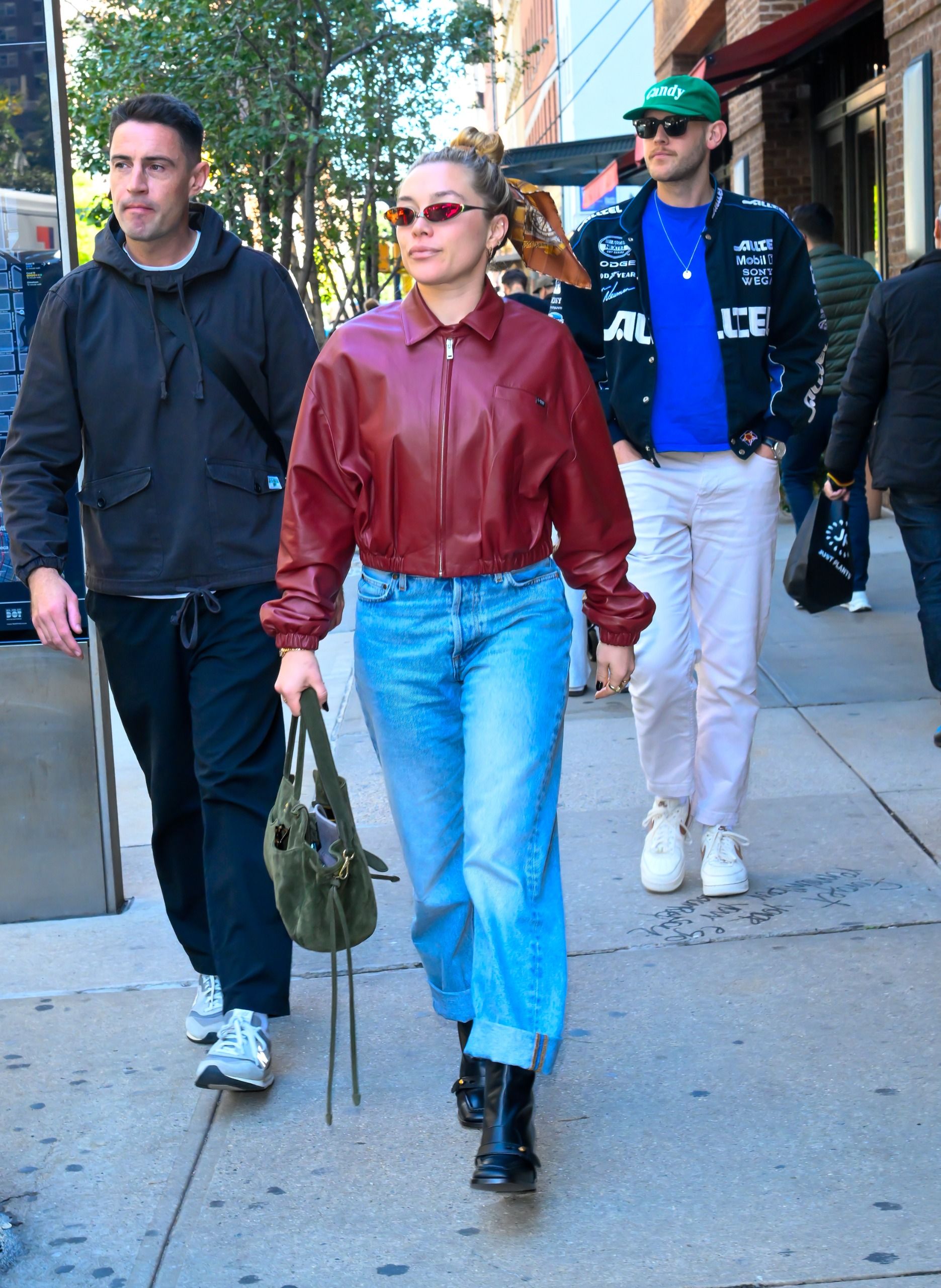 Florence Pugh walks down the street wearing a red leather bomber jacket with blue cuffed jeans and black boots.
