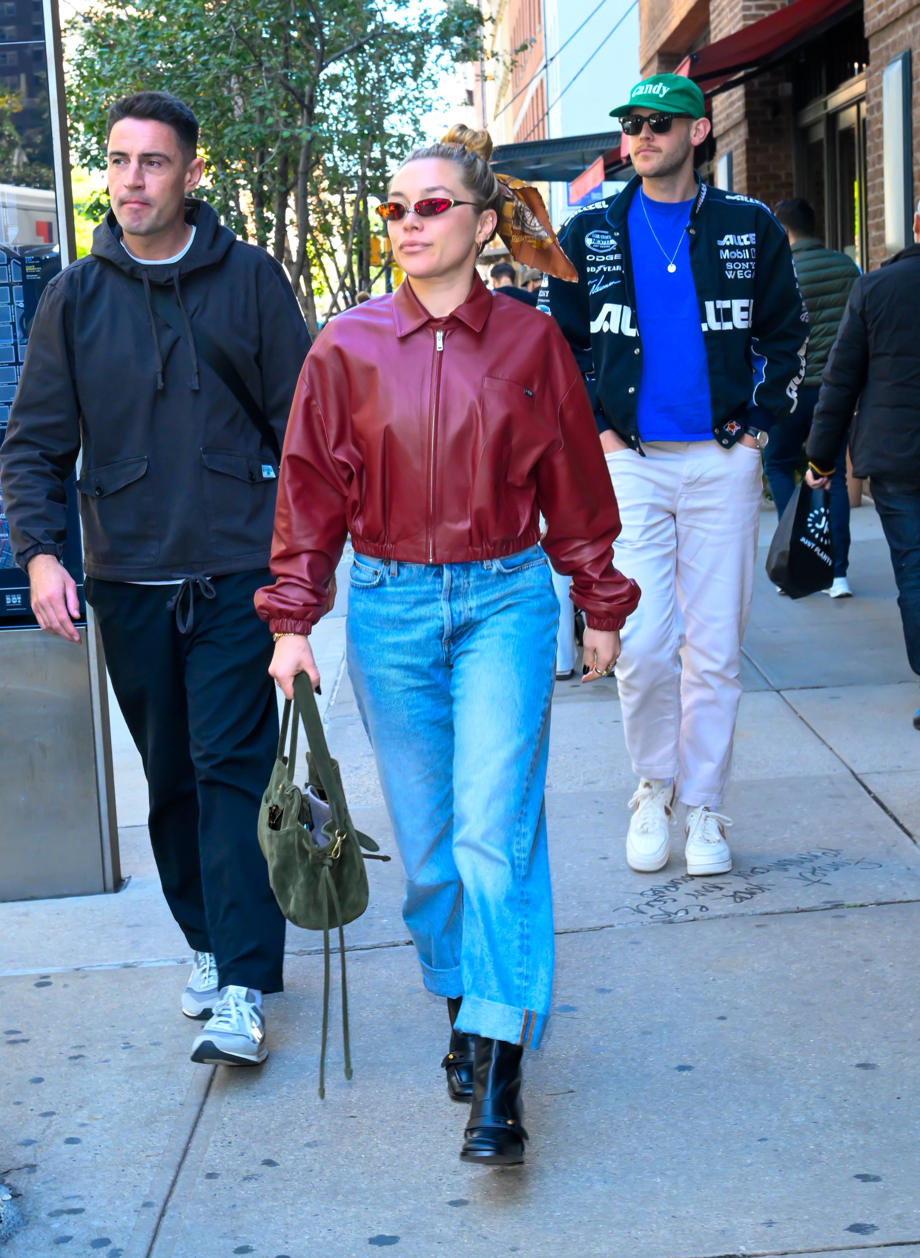 Florence Pugh walks down the street wearing a red leather bomber jacket with blue cuffed jeans and black boots.