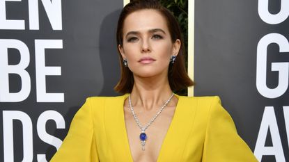 Zoey Deutch in a yellow jumpsuit with hands on hips wears a necklace with a large sapphire.