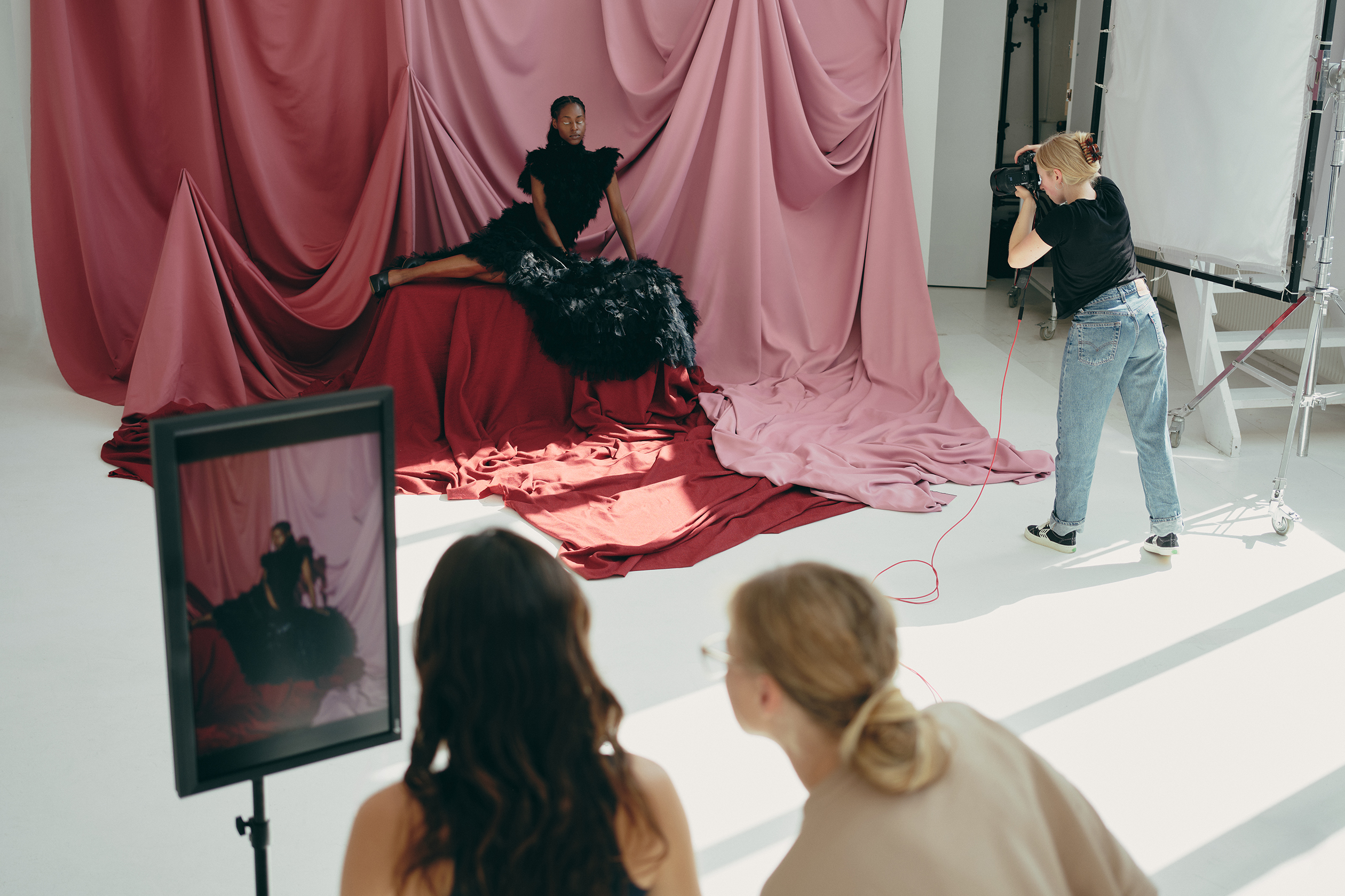 Fashion photoshoot in a studio with a model in a black feathered dress, a photographer taking pictures, and two people reviewing the shoot on a monitor