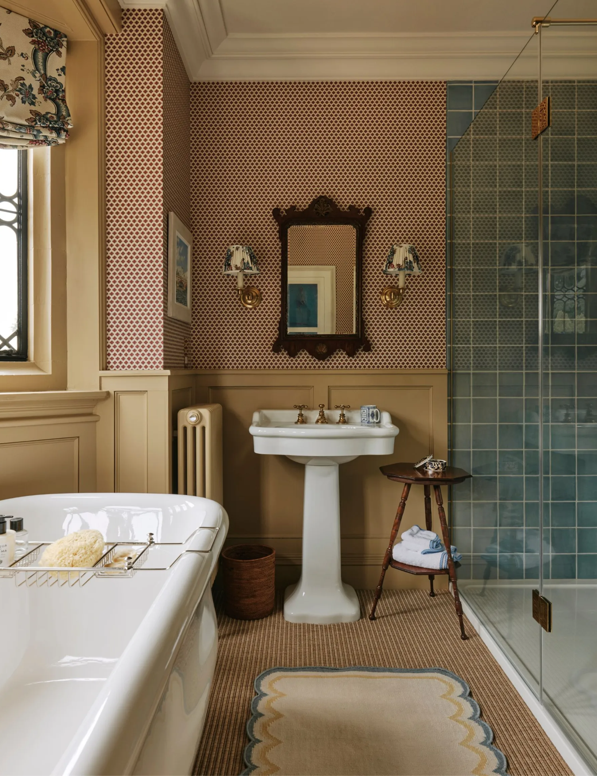 Bathroom with butter yellow panelling and window frame, polka dot wallpaper, blue tiled shower, jute carpet, pedestal sink and bath tub
