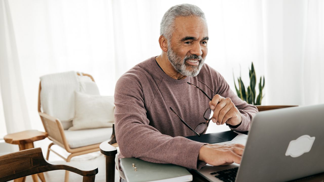 A retiree smiles while sitting at a table and looking at his laptop.