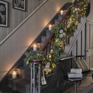 Wrapped gifts and pillow on wooden bench next to staircase that is decorated with natural greenery, fairy lights and sparkly baubles