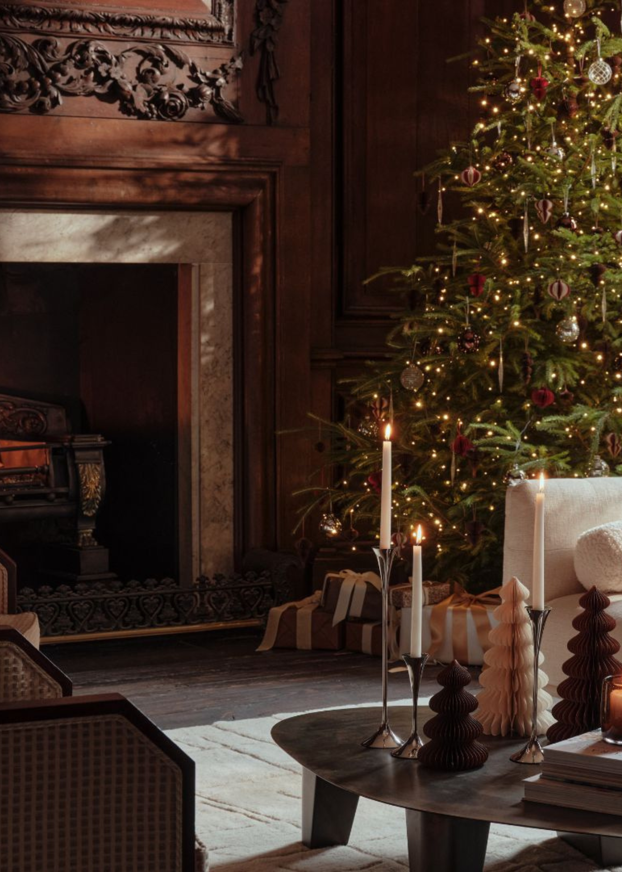 A living room with a Christmas tree by a fireplace with wooden engravings, a low coffee table with lit taper candles and paper Christmas trees