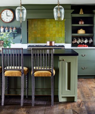Traditionally styled kitchen with dark wooden flooring, green painted walls and cabinets, bright tiled green wall above stove, kitchen island with black granite countertop, two wooden bar chairs with orange seat upholstery, two glass pendants hanging over island