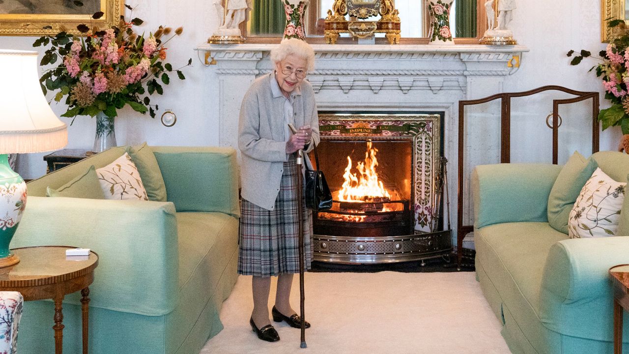 Queen Elizabeth wearing a plaid skirt and cardigan holding a can in front of a fireplace 