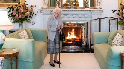 Queen Elizabeth wearing a plaid skirt and cardigan holding a can in front of a fireplace 