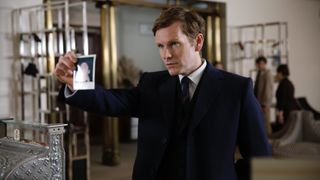 Shaun Evans in a dark suit as Endeavour holds up a photo in Endeavour.