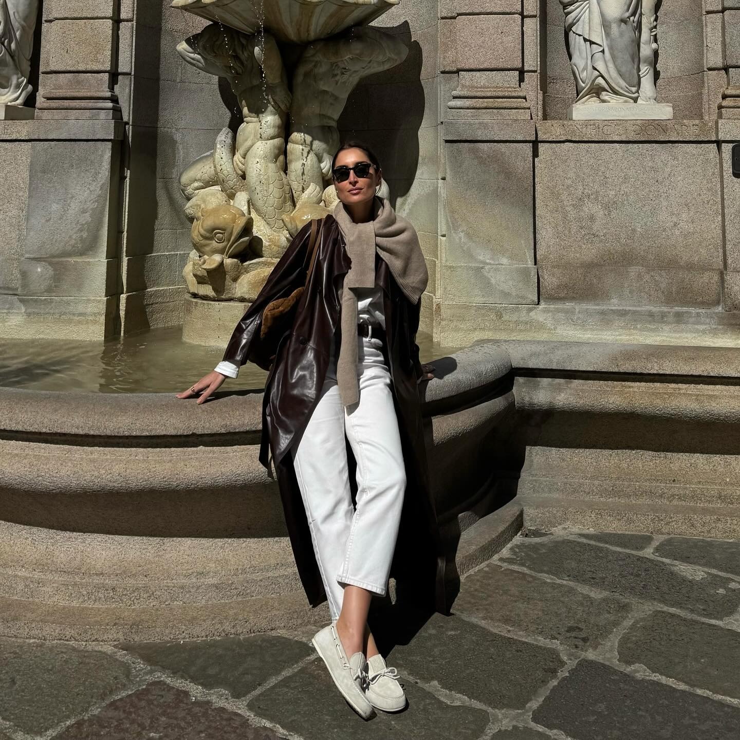 Fashion person Erin shows how to style white jeans. She pairs her jeans with a vinyl leather trench, a cashmere jumper, a suede bag, a black belt and suede boat shoes. She is standing in front of a fountain in Mila, looking directly into the camera in a recent image from her Instagram.