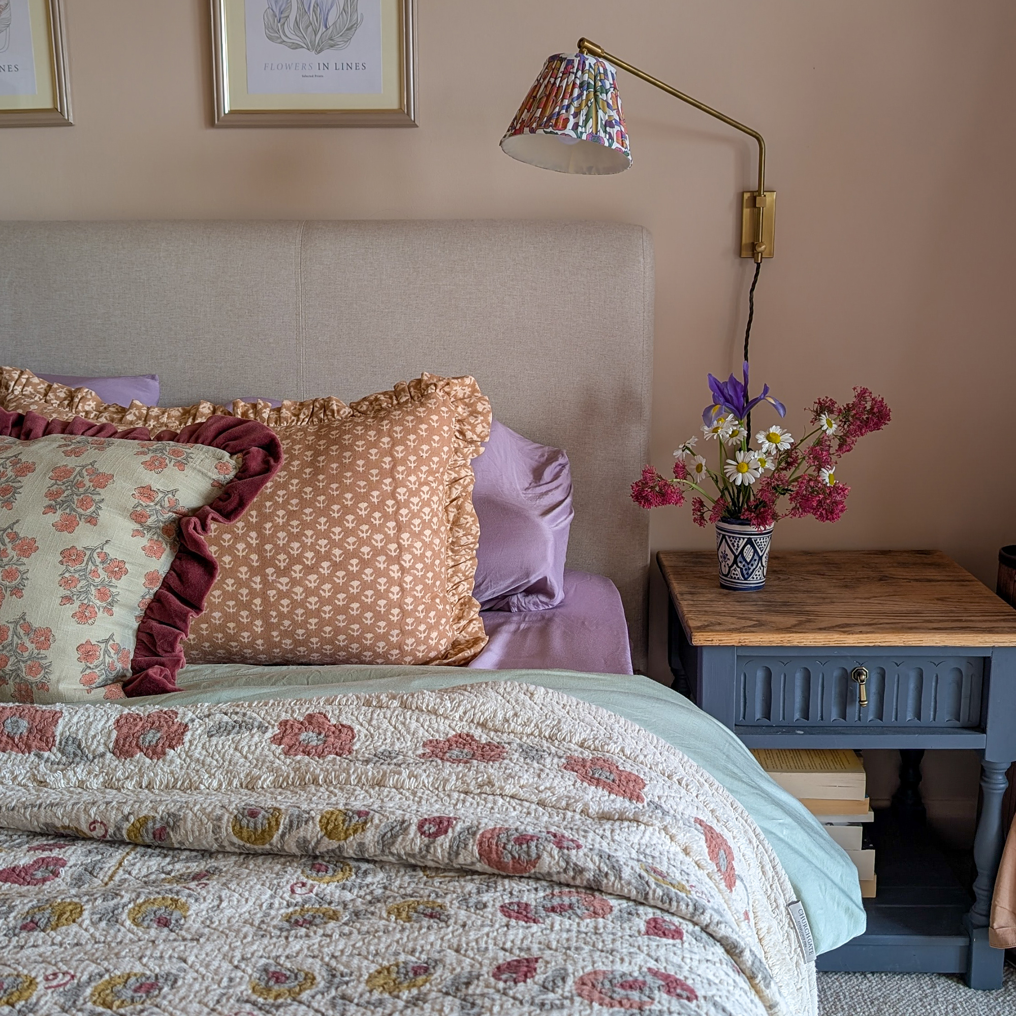 Pale pink bedroom with navy painted side table and beige headboard and a bed dressed with floral quilt and an assortment of patterned cushions
