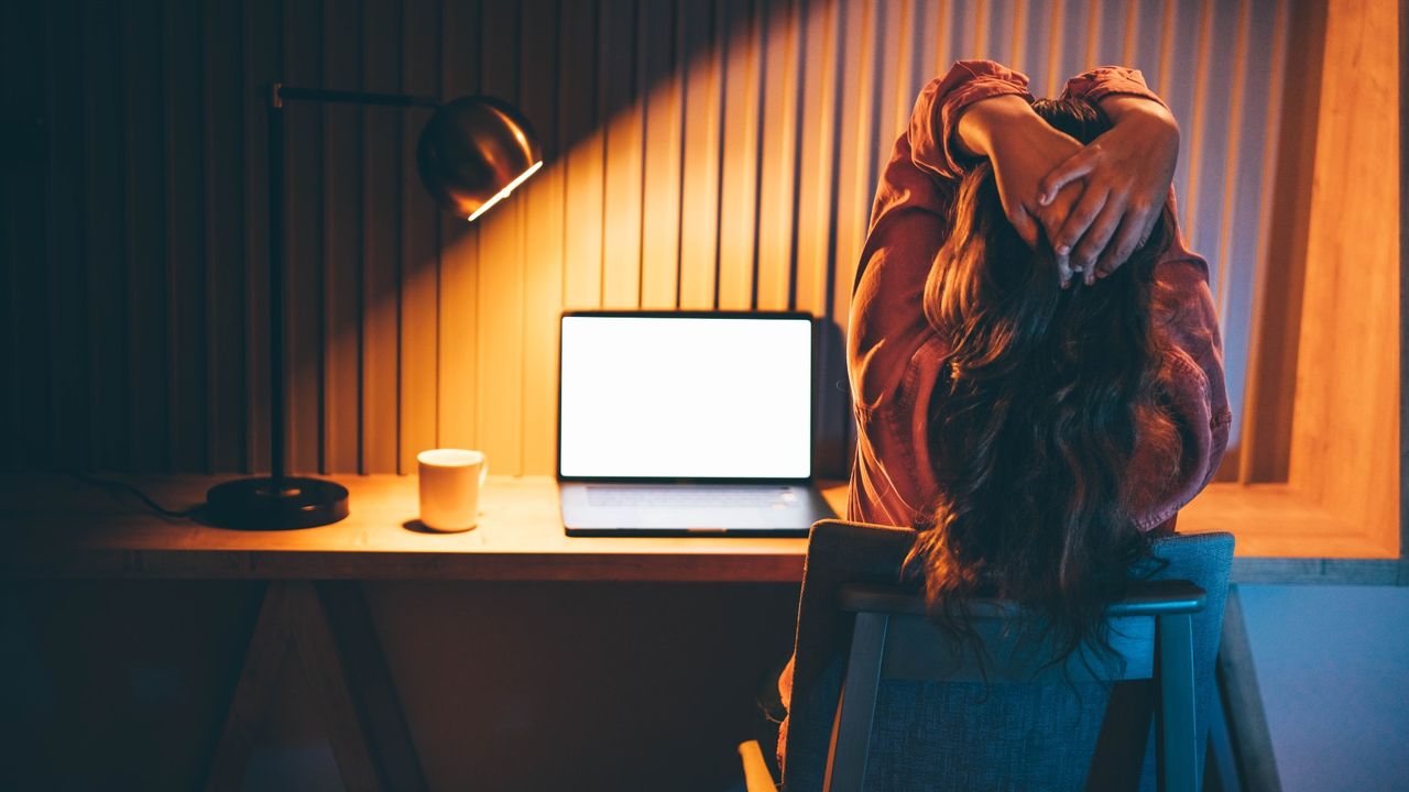 A frustrated freelancer sits at her desk in front of her laptop, in a dark office. 