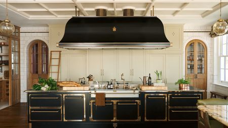 A traditional kitchen with cream cabinets with cut outs at the top, a wood and glass walk-in pantry, and a black island with brass cabinet borders