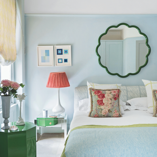 Master bedroom with cream carpet, pale blue walls and double bed with light blue blanket and vintage cushion with floral pattern, lamp with red lampshade on mirrored beside table.