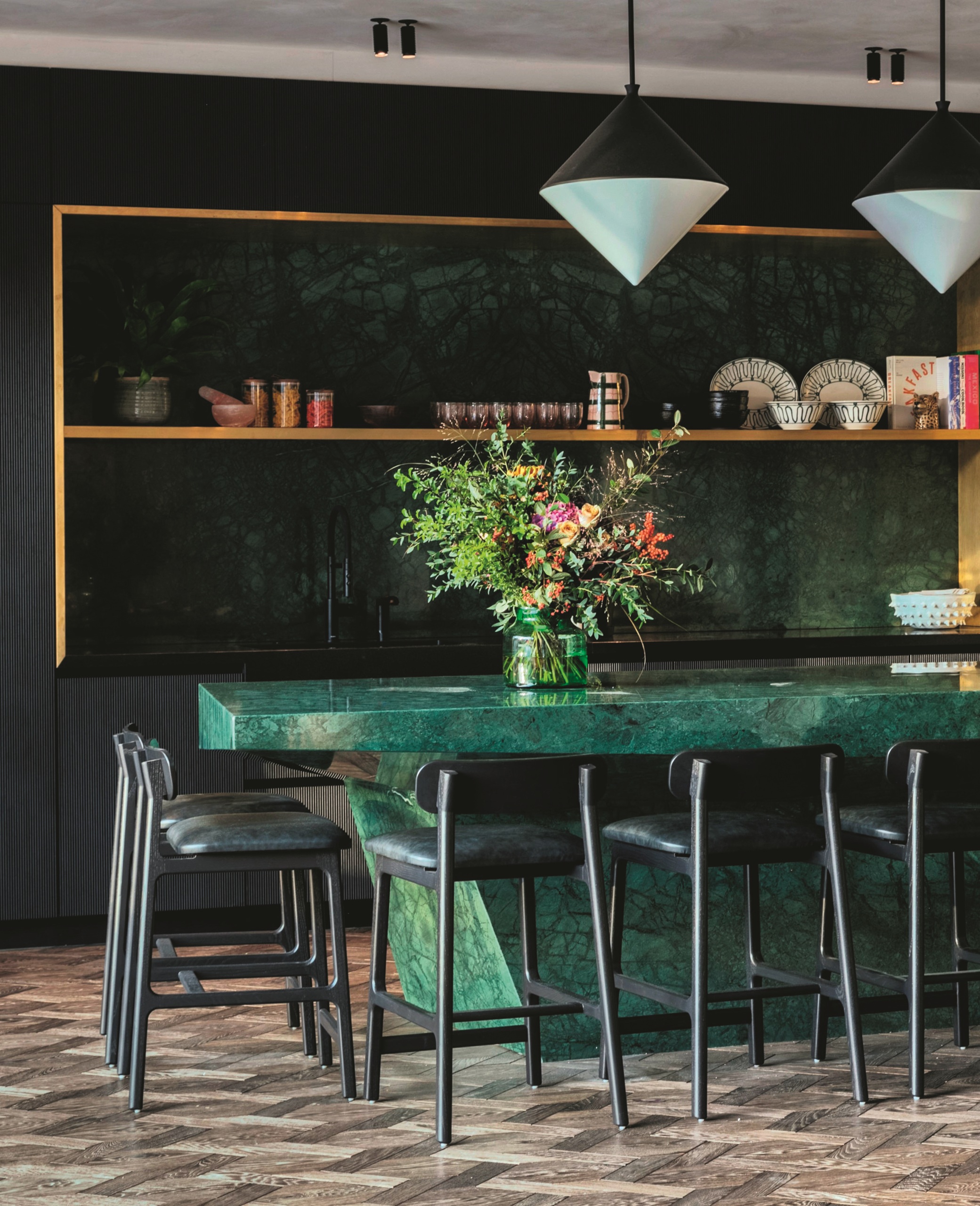 green marble kitchen with angled island, timber floors, black bar stools, green backsplash and black fluted cabinetry with gold trim
