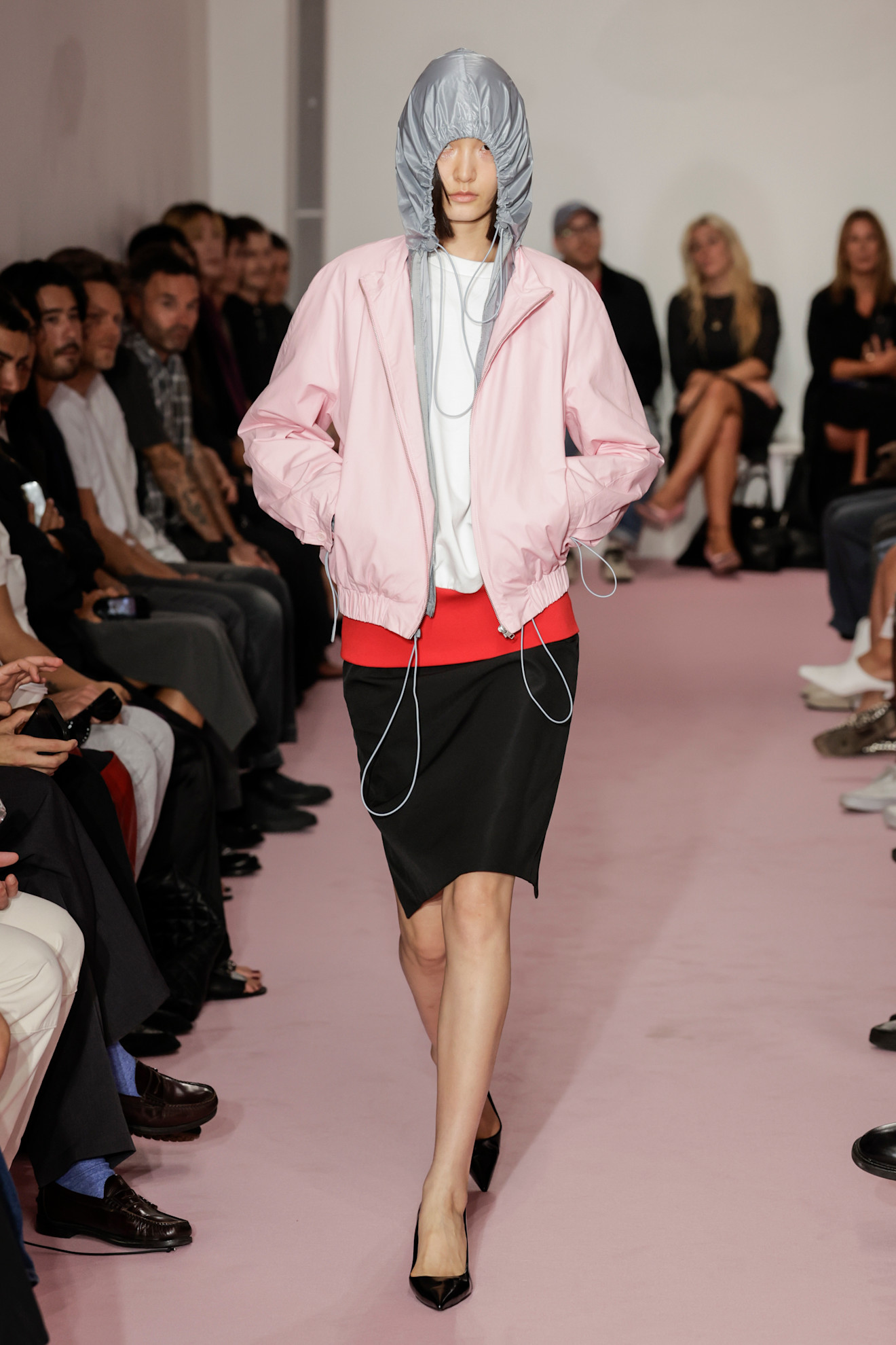 Layering staples for spring 2026 are shown in a photo from LII's runway show, with a model walking while wearing a pink parka jacket styled on top of an open gray hoodie with a white t-shirt peeking out underneath styled with a black and red two-tone pencil skirt and black pointed heels