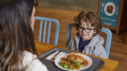 A smiling child sits at a table with a parent and a plate of warm food