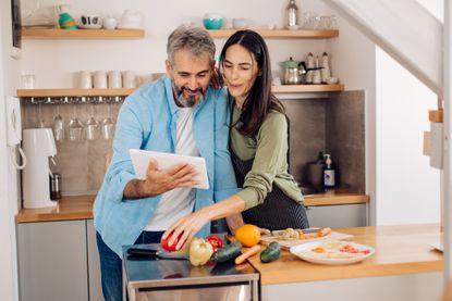 Mature couple using tablet and preparing a takeaway vegetable meal at their kitchen.