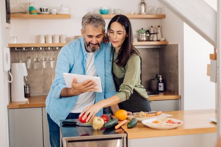 Mature couple using tablet and preparing a takeaway vegetable meal at their kitchen.