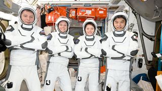 Two men and two women, all wearing white pressure suits and helmets, pose for a photo together inside a space station module
