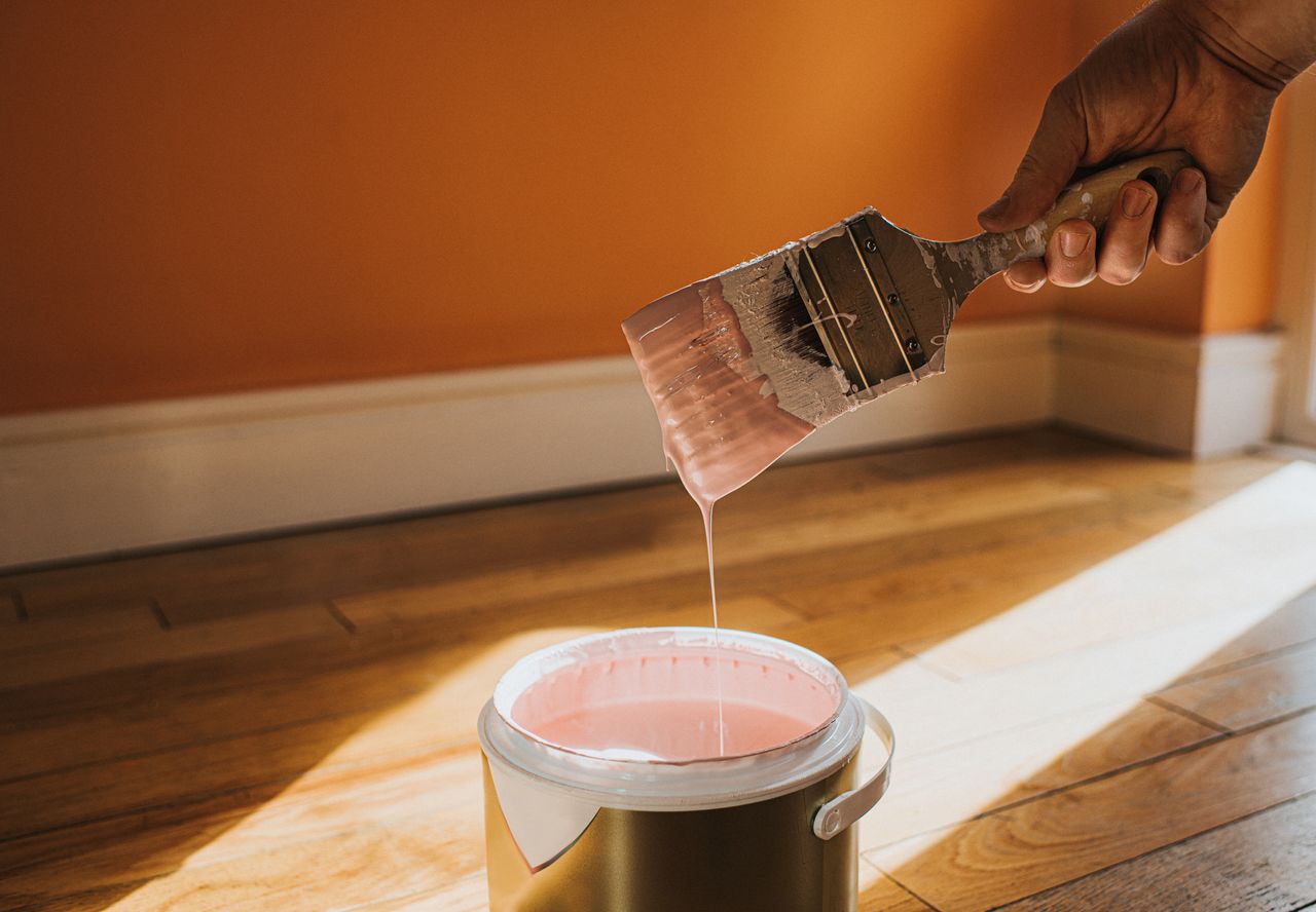 Hand dipping a paint brush into a large tin of pink paint. 