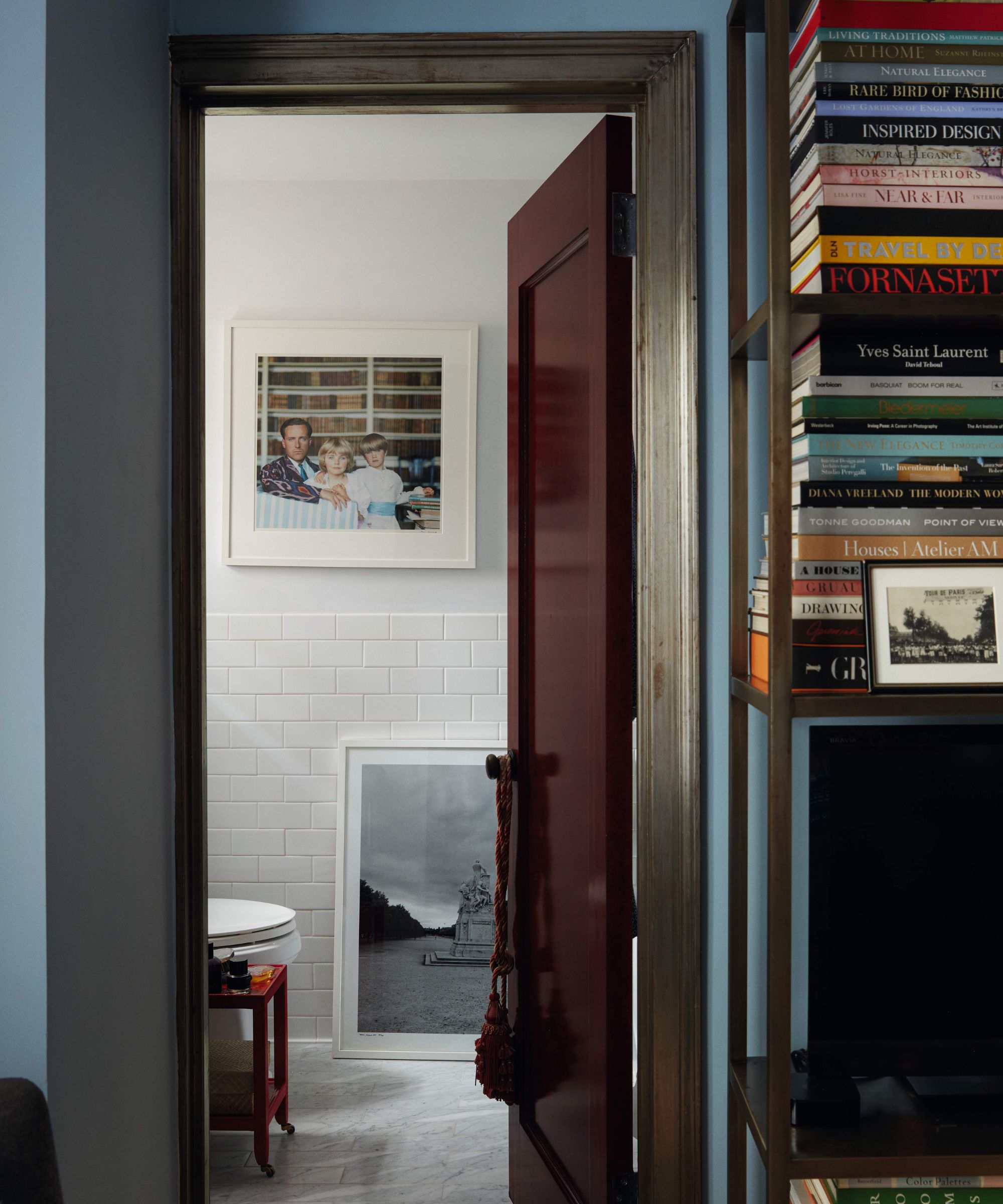 A view through a doorway into a white-tiled bathroom featuring a marble floor, a large framed family portrait, and a black-and-white landscape photograph leaning against the wall