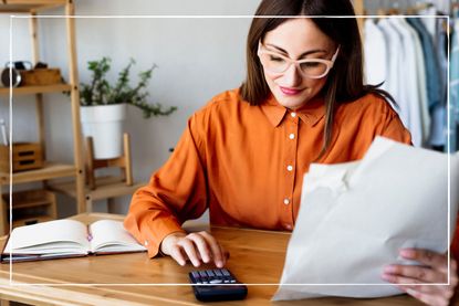 Woman in orange shirt, wearing glasses, sitting at a desk at home, holding pieces of paper and using a calculator