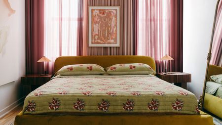 An fabric-covered bed with a green bedspread, backdropped by curtains and a hanging painting