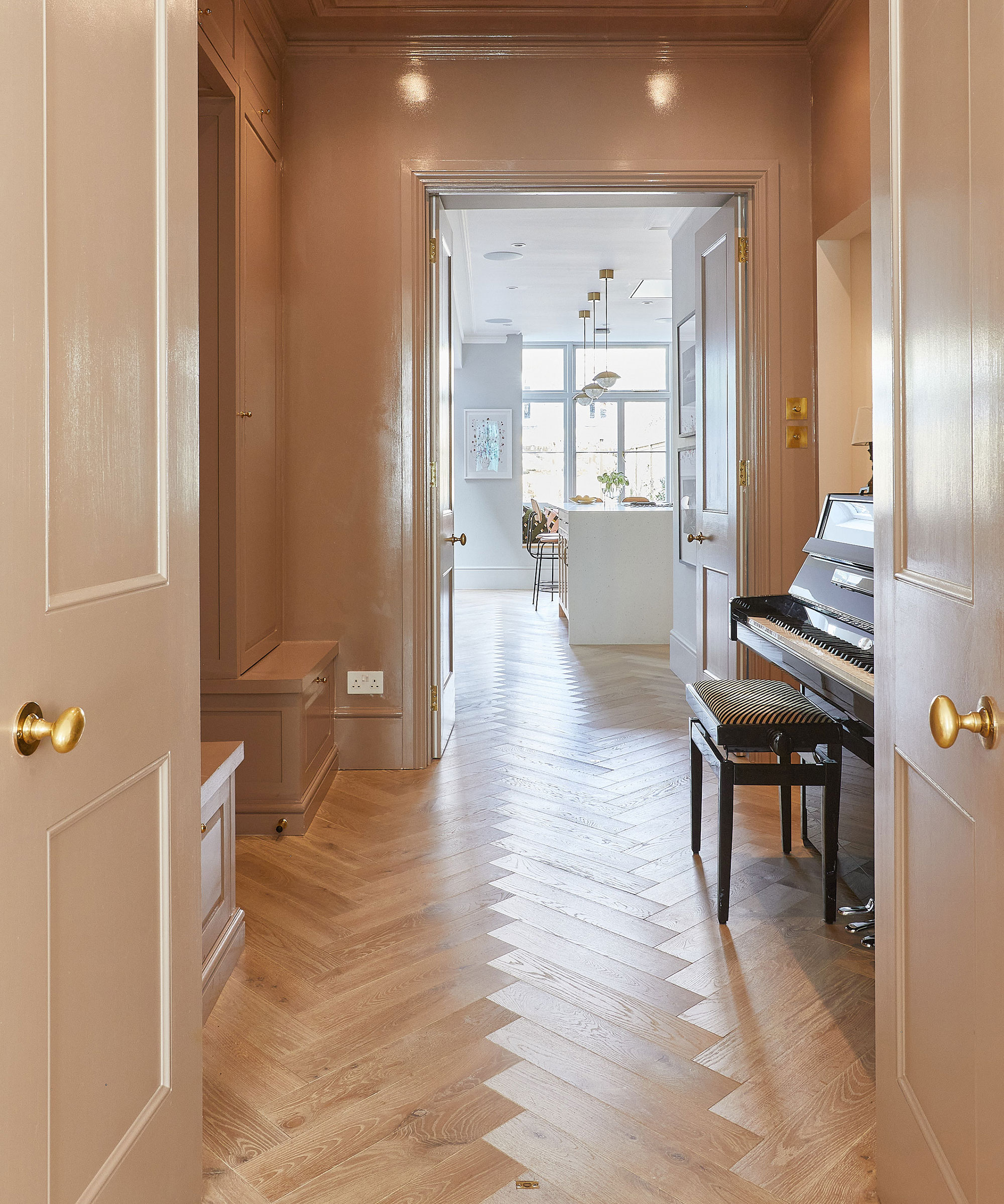 classic entrance hallway with herringbone wooden flooring and piano leading to white kitchen beyond