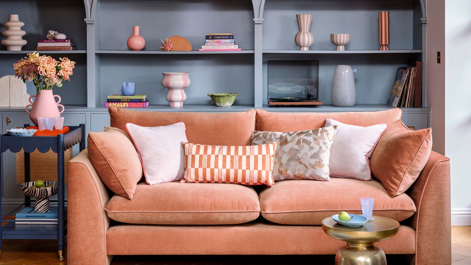 A plush orange sofa with white and orange throw cushions, in front of blue built-in bookshelves, decorated with small vases and books. 