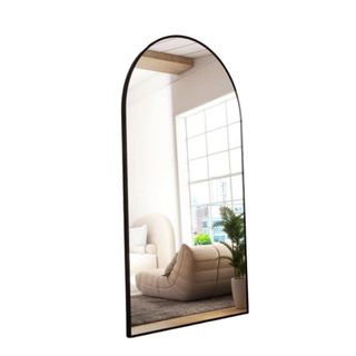 An arched full-length black mirror tilted to the right reflecting a gray living room with a seat, plant, and window