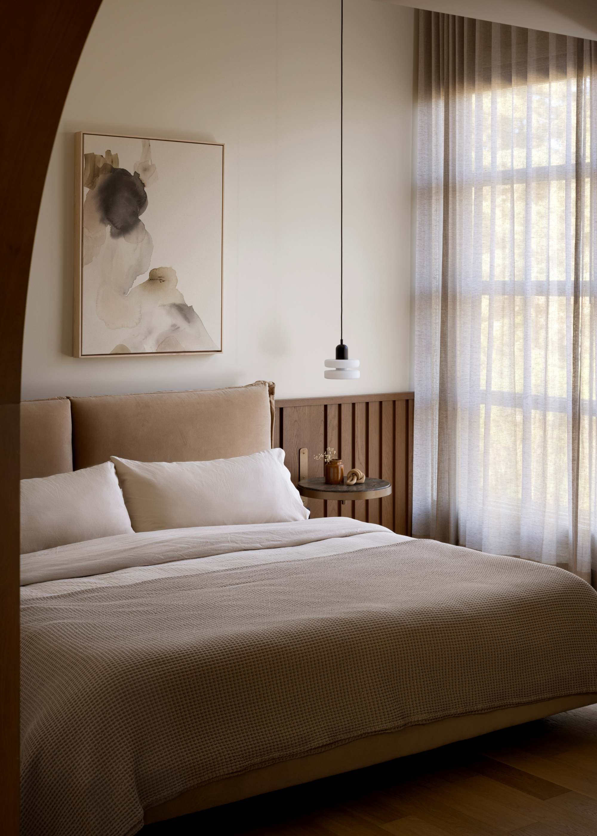 A white bedroom with a beige bedscape, sheer window treatments, a painting above the bed, a floating nightstand with a pendant lamp