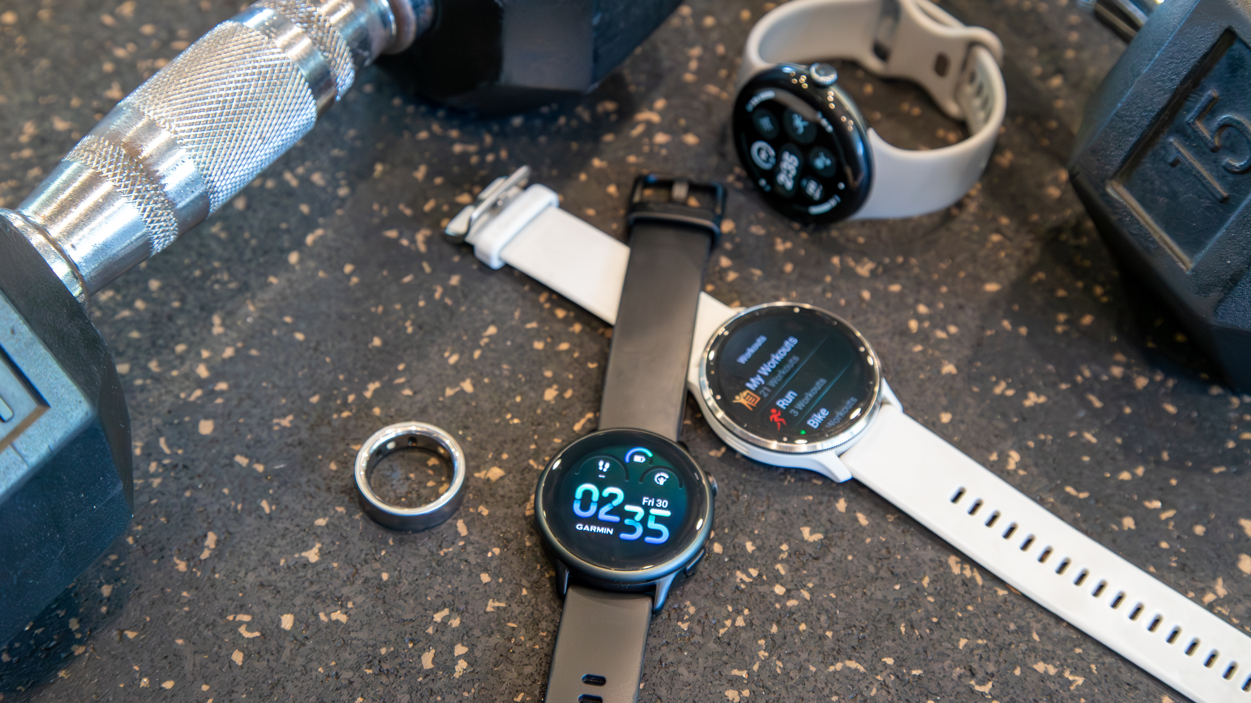 The Garmin Vivoactive 6, Venu 3, Pixel Watch 3, and Oura Ring 4 next to dumbbells