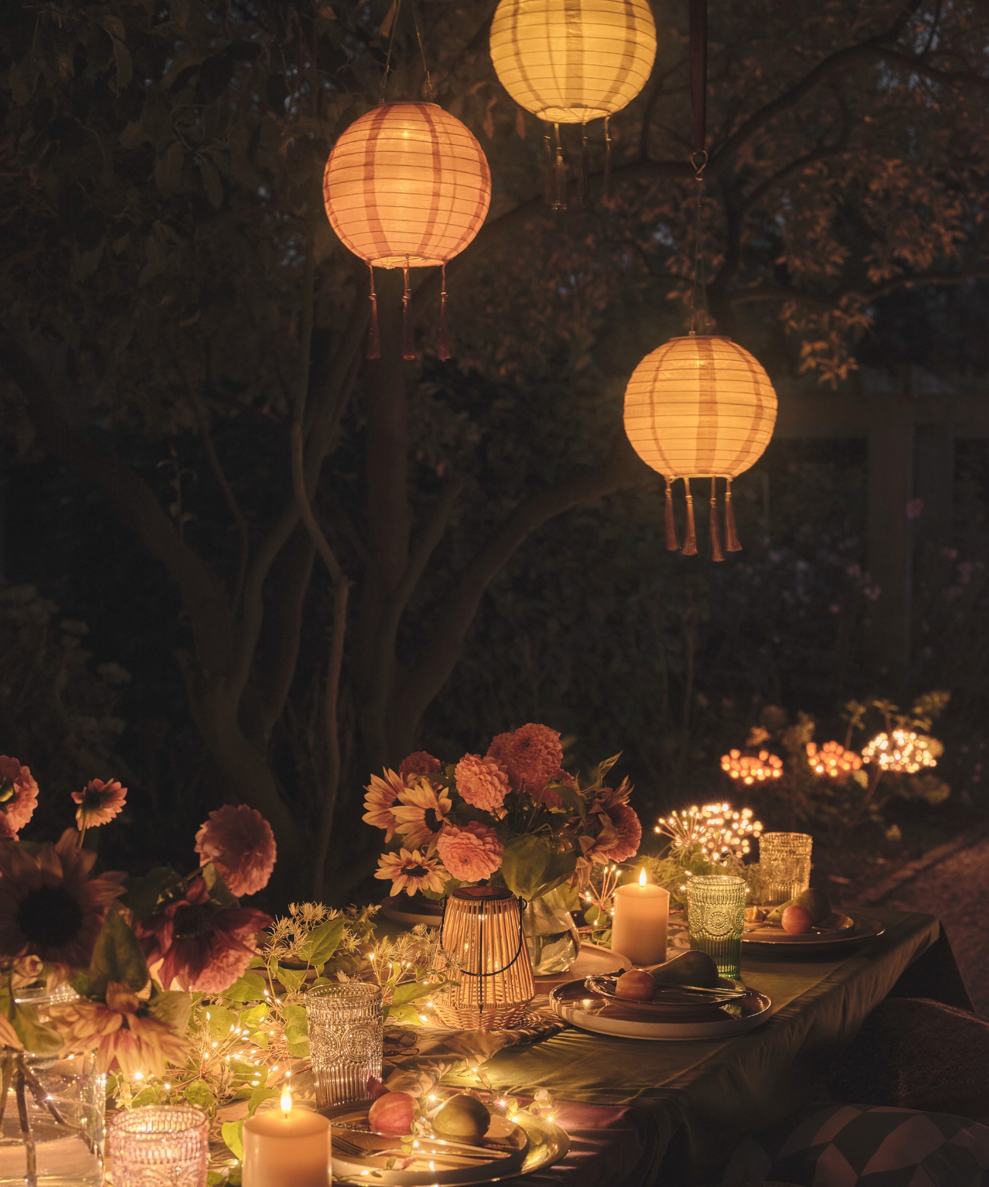 A garden after dark with a set dining table decorated with flowers and lights and three hanging lanterns above it