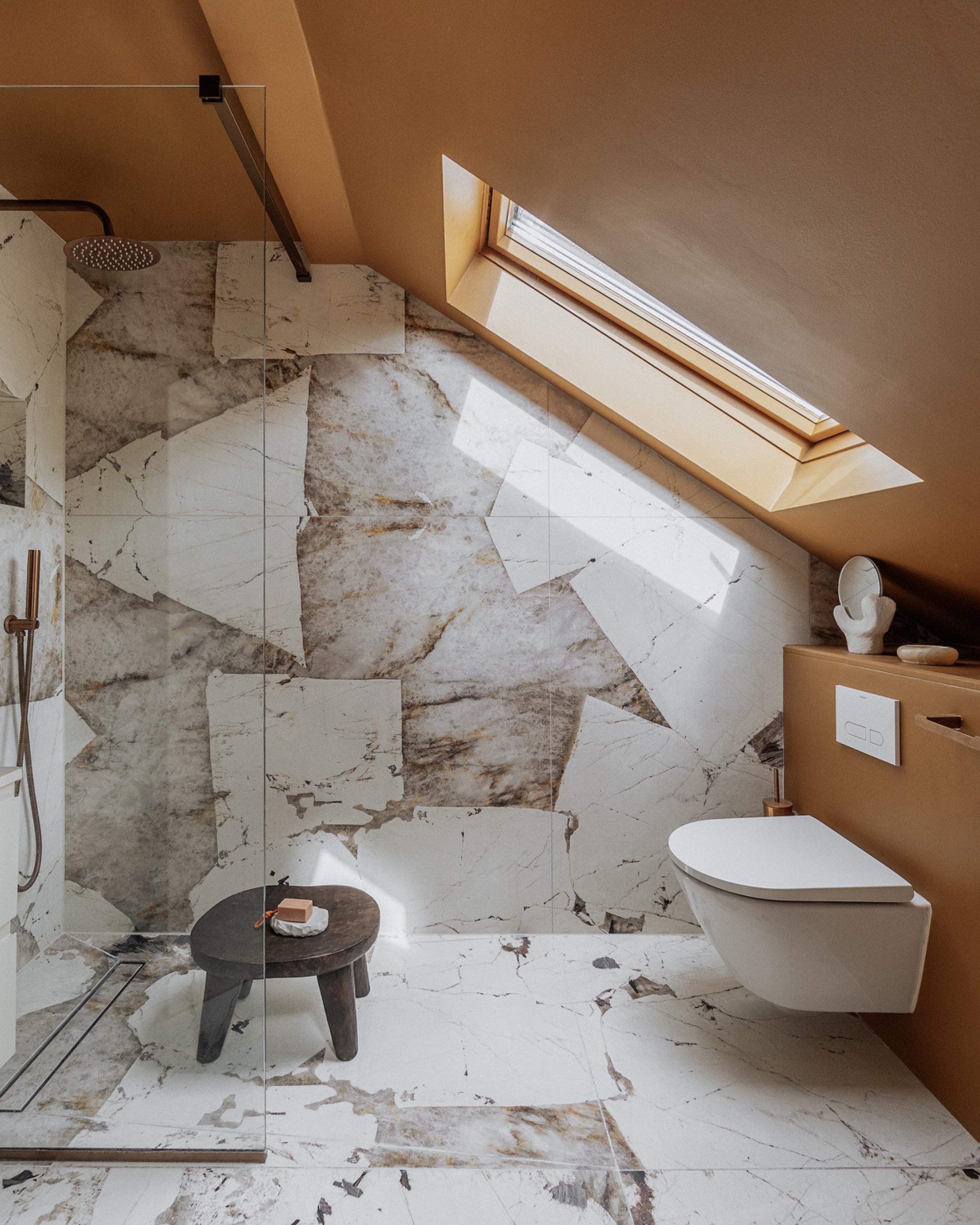 Bathroom beneath a sloped ceiling and skylight, with veined stone walls and flooring, a glass shower screen and a tile-in linear drain along the shower&amp;rsquo;s edge.