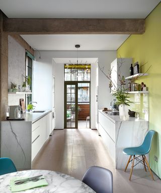 A marble galley kitchen with yellow wall, open shelving and teal chairs.