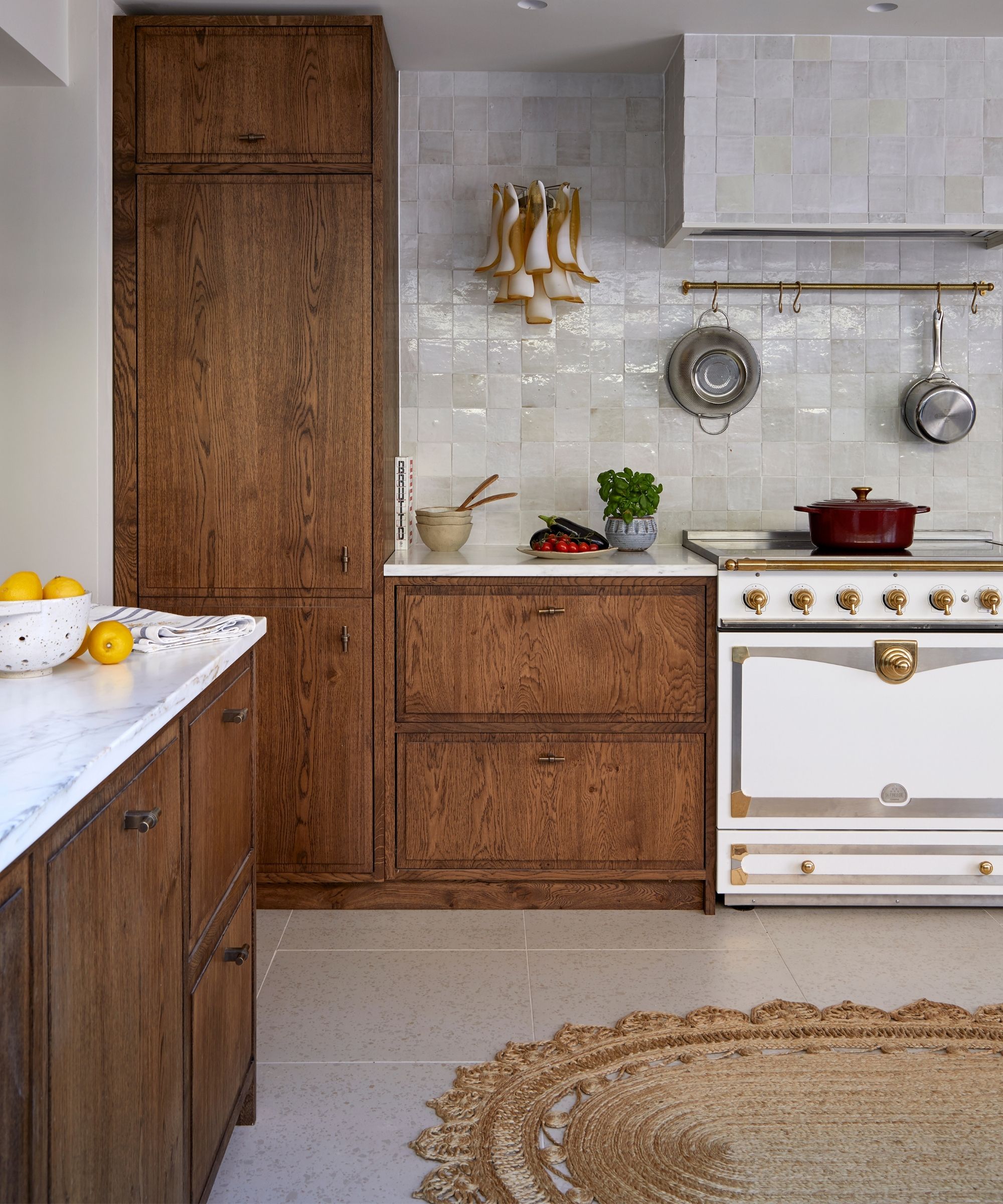 A white kitchen with white zellige tiles, brown cabinetry, a white stove, white marble countertops, light tile flooring, and a large oval jute rug.