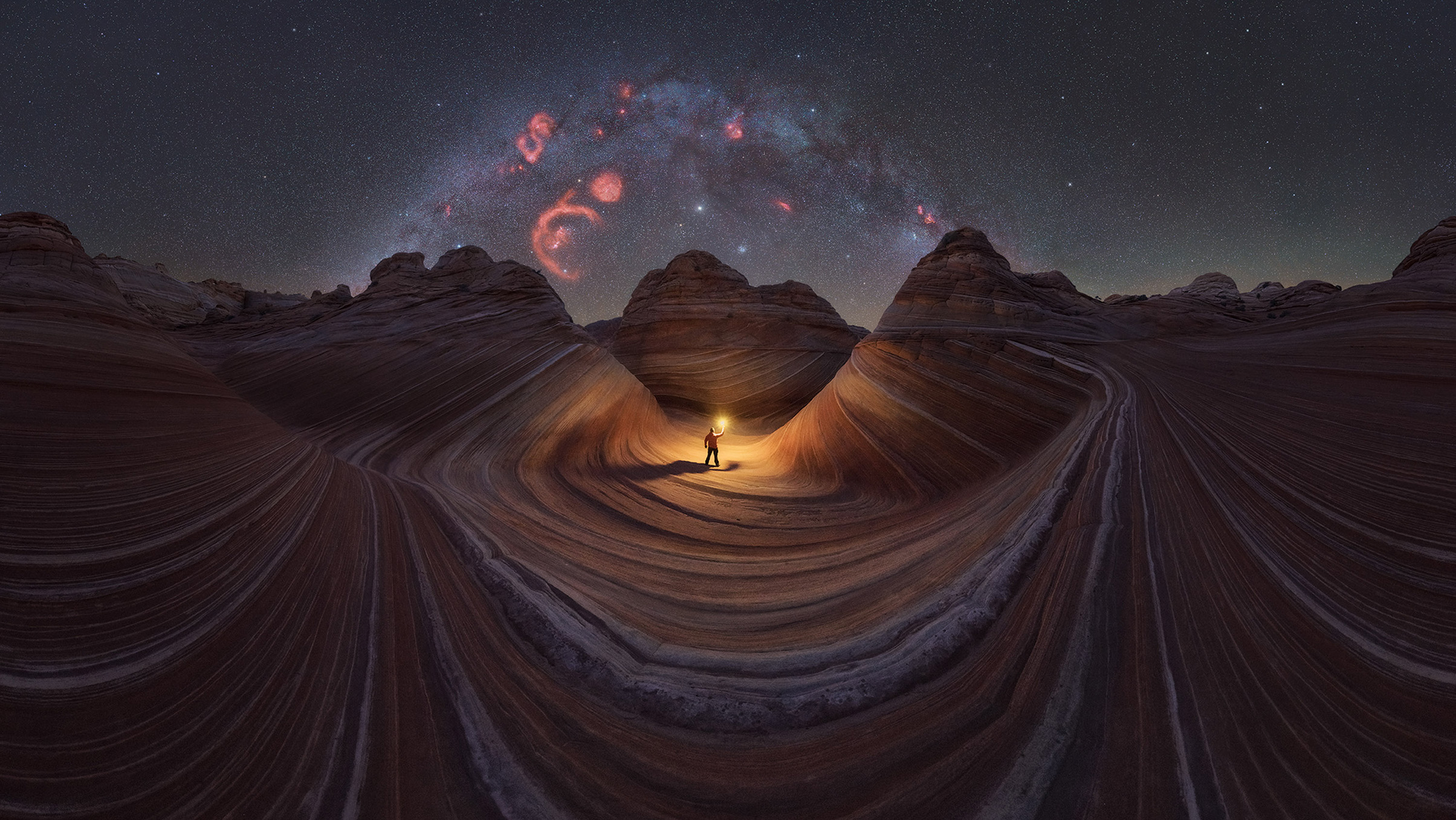 A person stands with a glowing lantern in the winding, colorful rock formations under a starry sky with a vibrant galaxy arc above