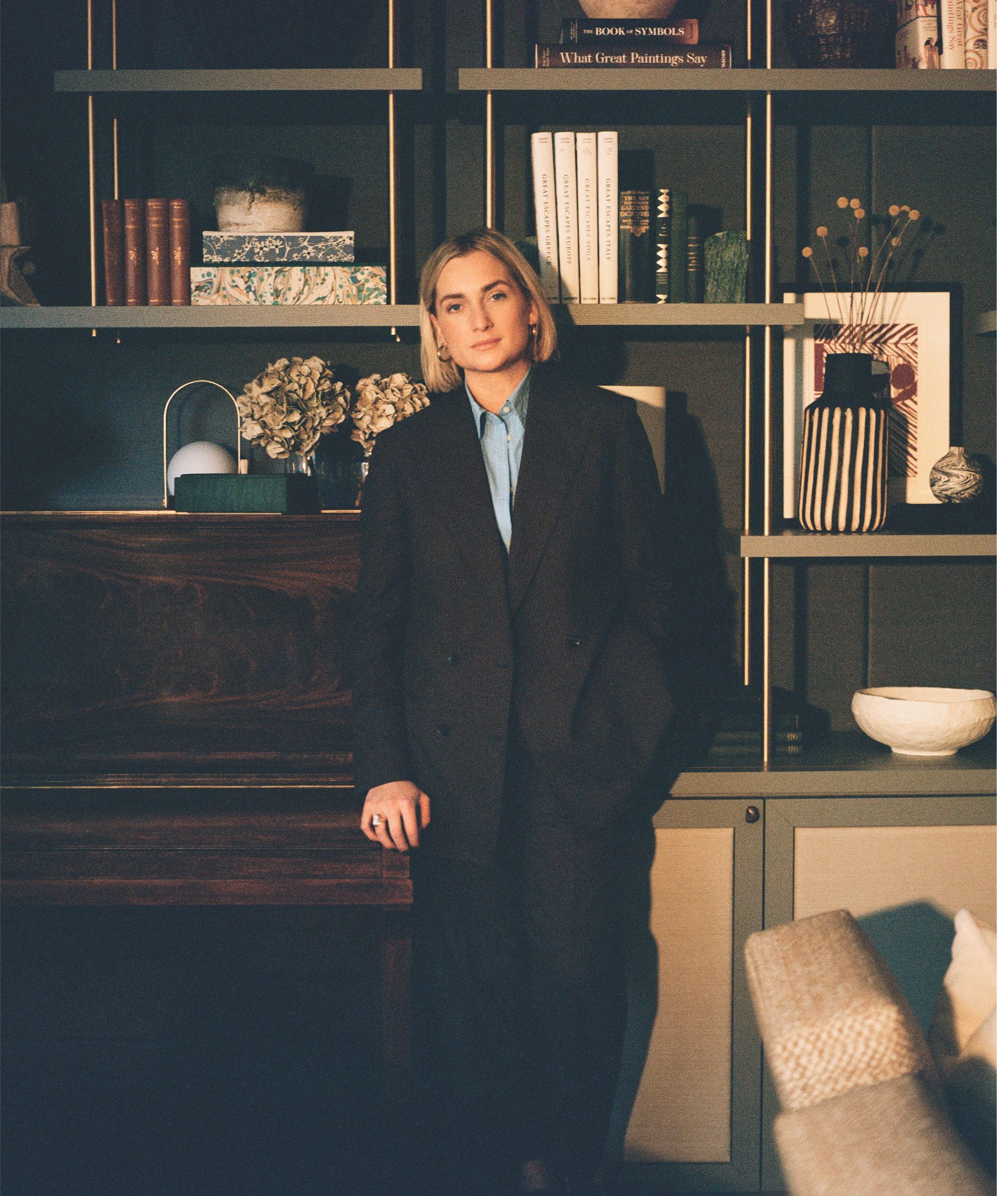 A woman with short blonde hair in a black blazer suit leaning against shelves